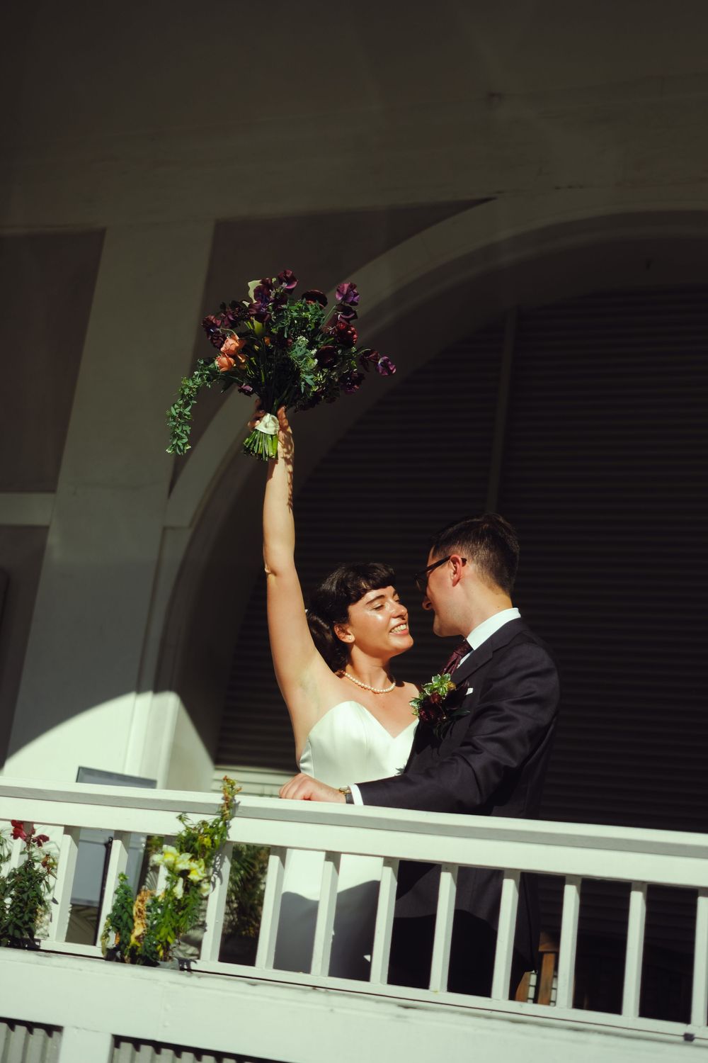 Joyful bride raises bouquet triumphantly on a white balcony while groom embraces her after wedding ceremony.