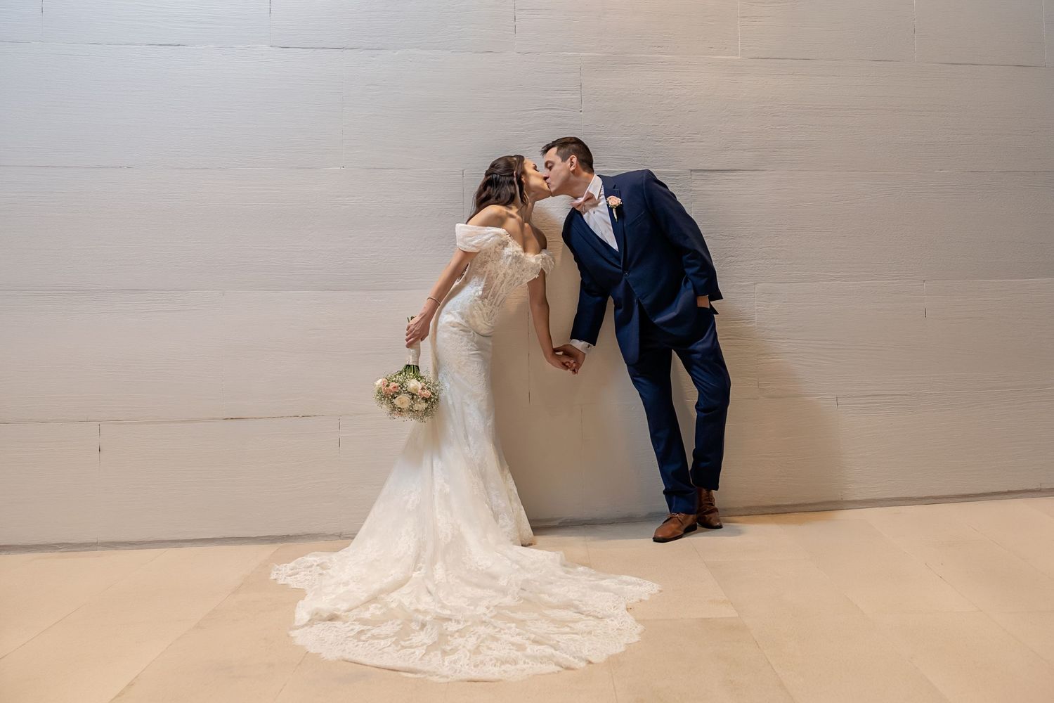 Bride in white gown and groom in navy suit share a romantic kiss against a white wall at their wedding.