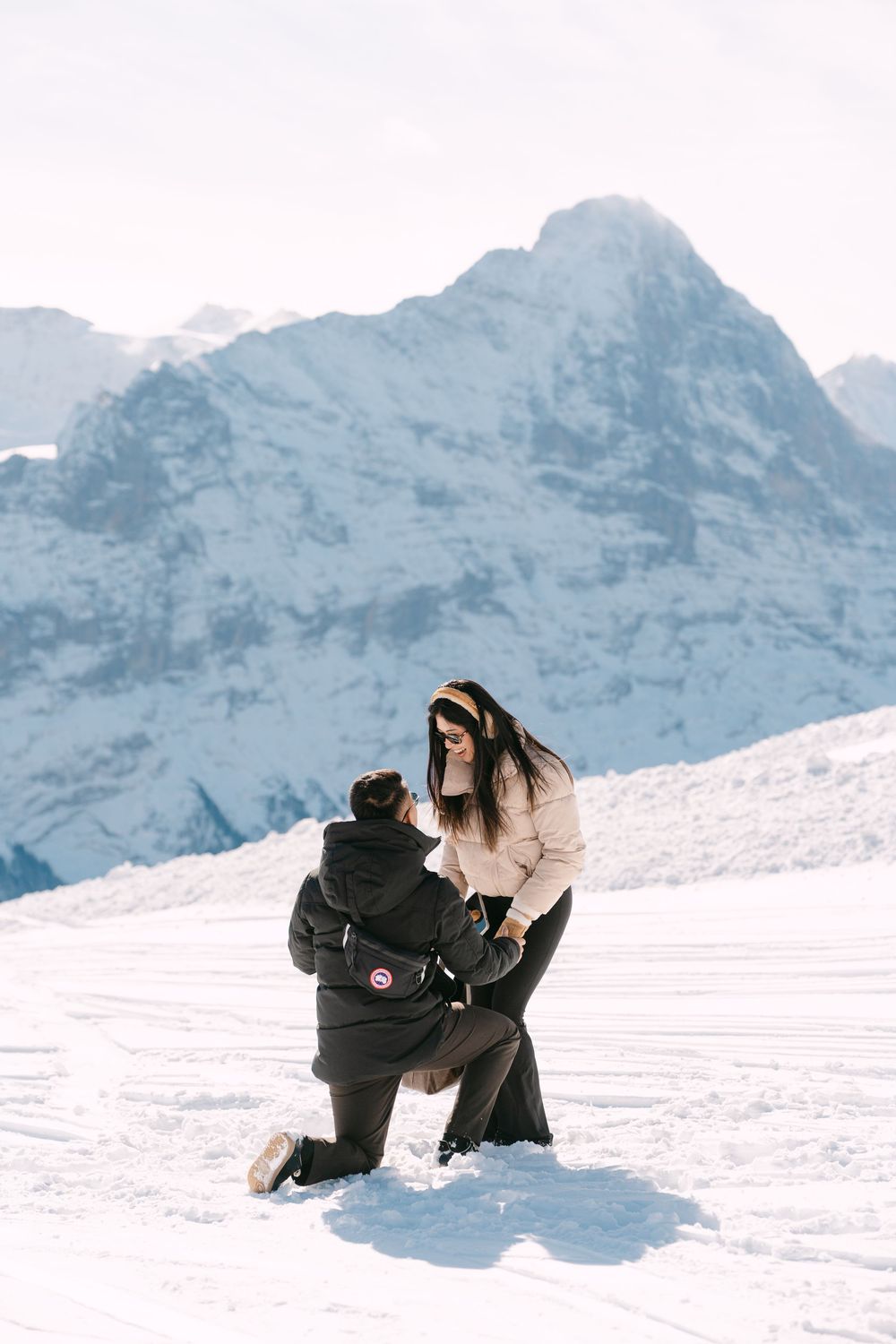Two people in winter coats share a romantic moment on snowy mountain slopes with majestic peaks in the background.
