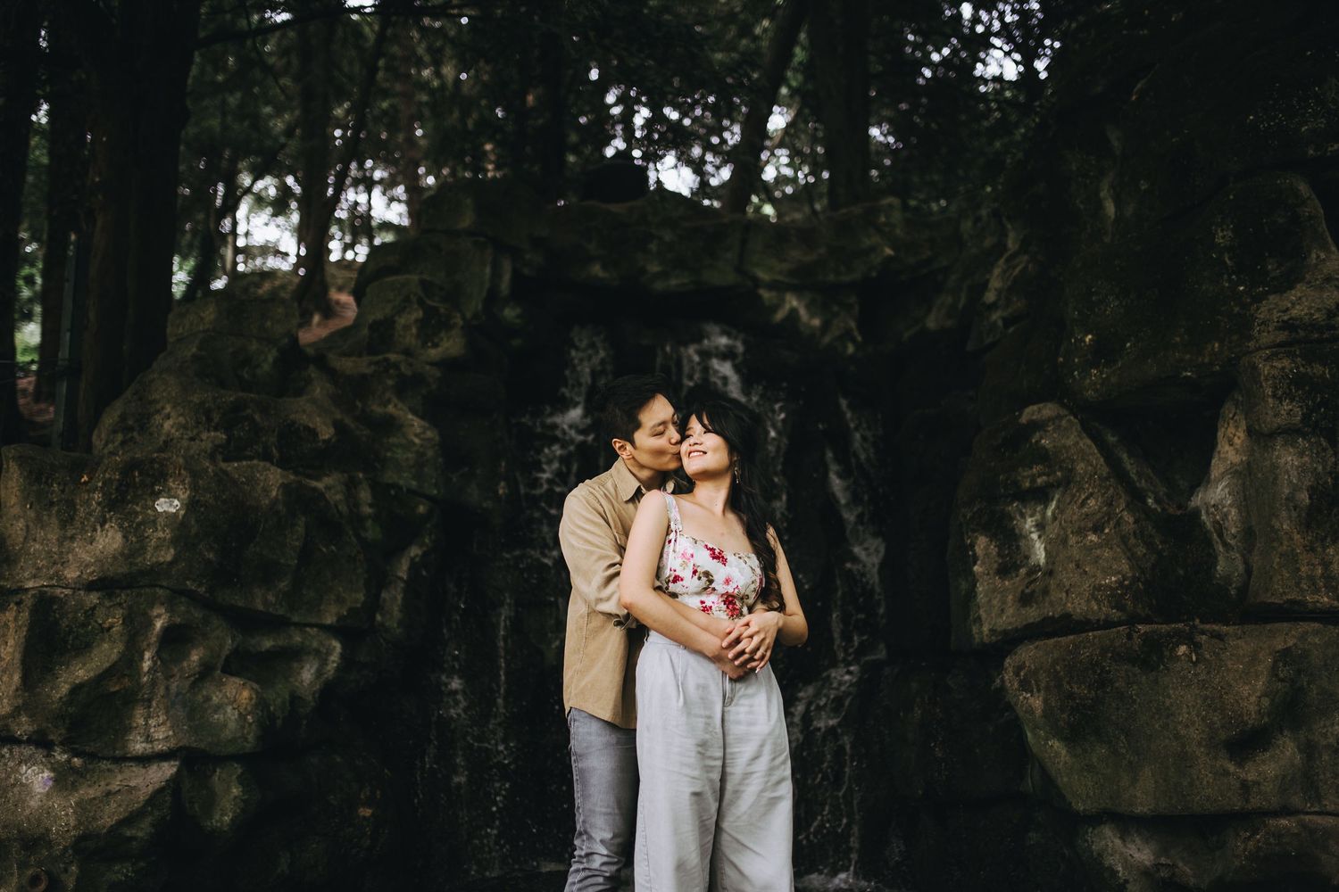Couple embraces against dark rocky backdrop in moody outdoor setting.