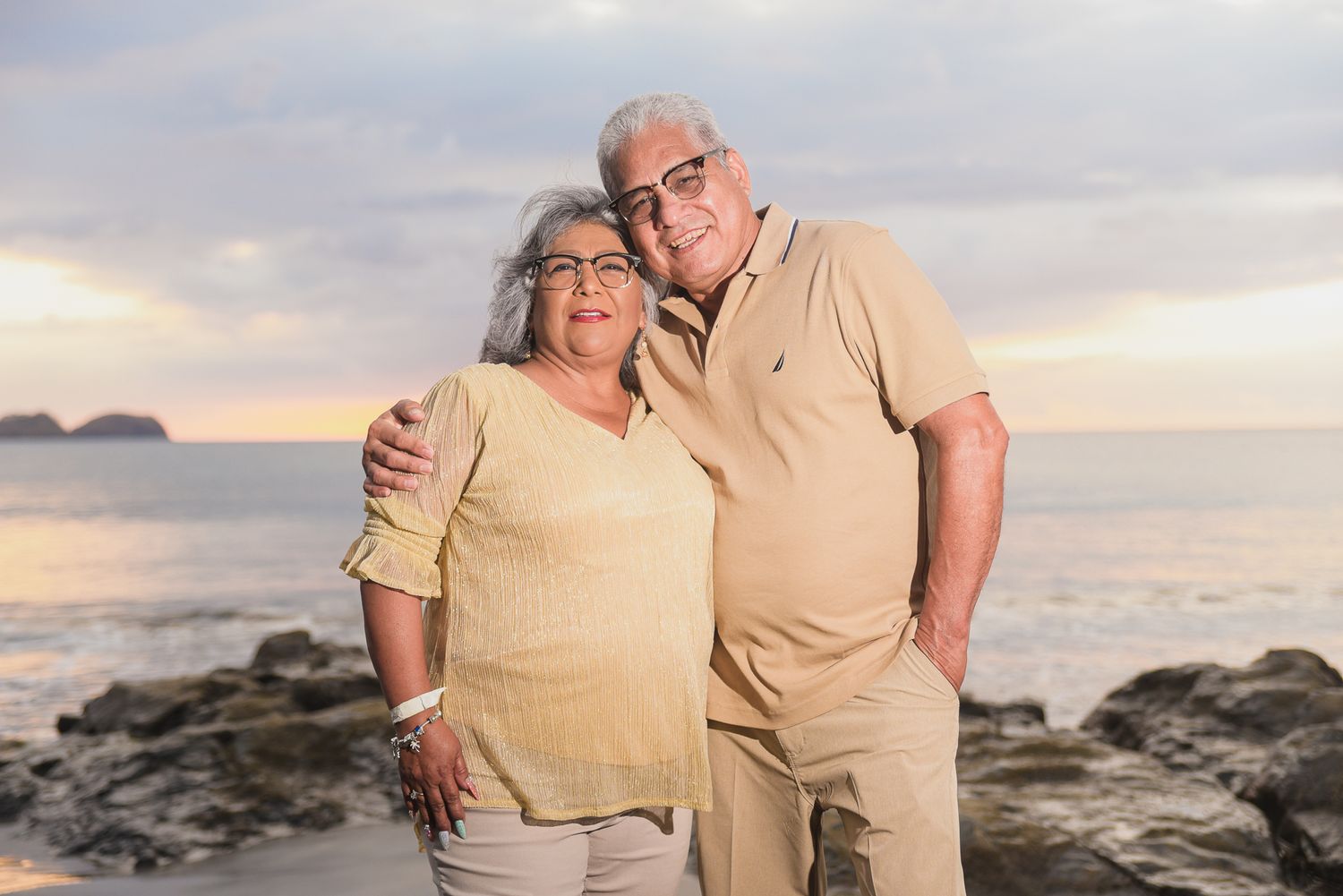 Couple session photo, in casual attire shares tender moments on a rocky beach at sunset. Riu Guanacaste, Costa Rica