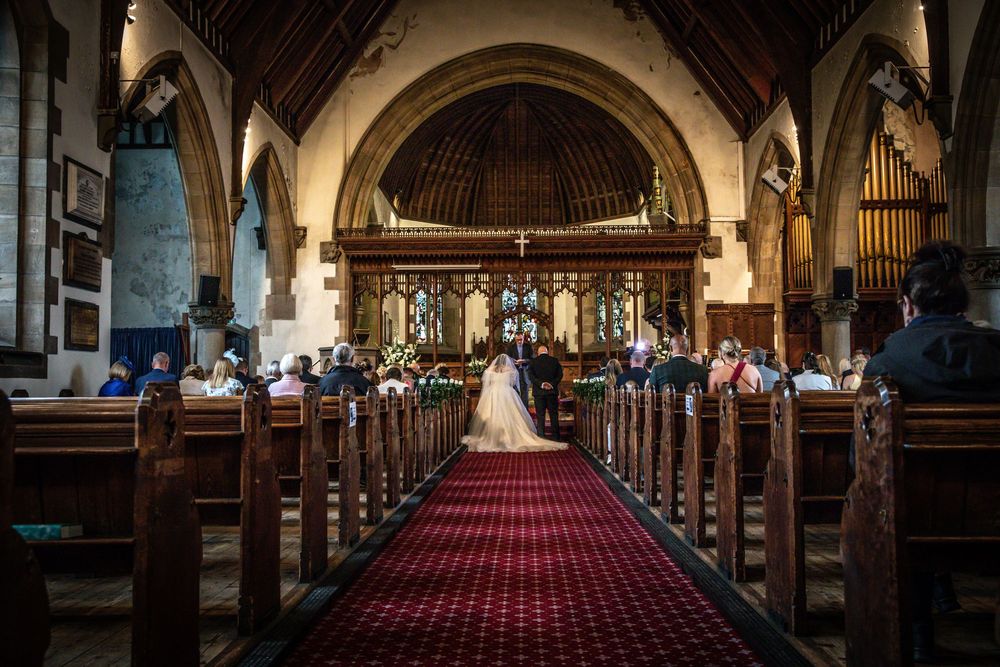 Bride walks down a red-carpeted church aisle lined with wooden pews filled with wedding guests during ceremony.