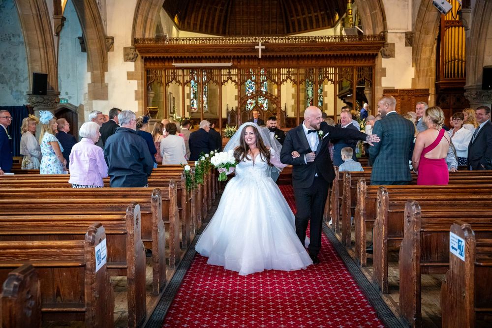 Bride in a full ballgown walks up the church aisle with groom after wedding ceremony on red carpet.