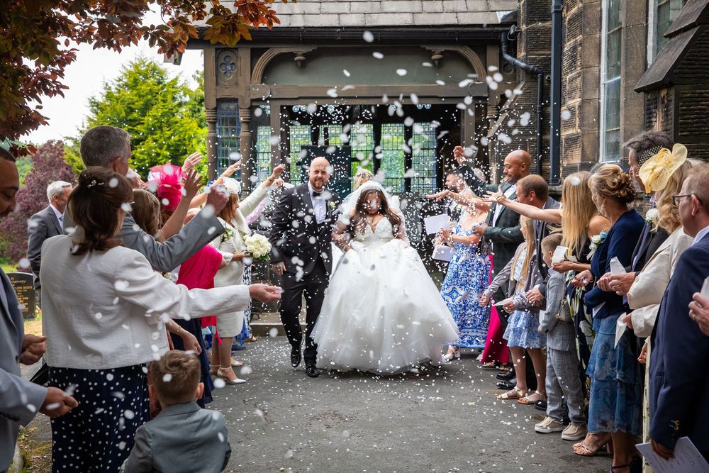 Newlyweds exit church entrance as guests shower them with white confetti in joyful outdoor celebration.