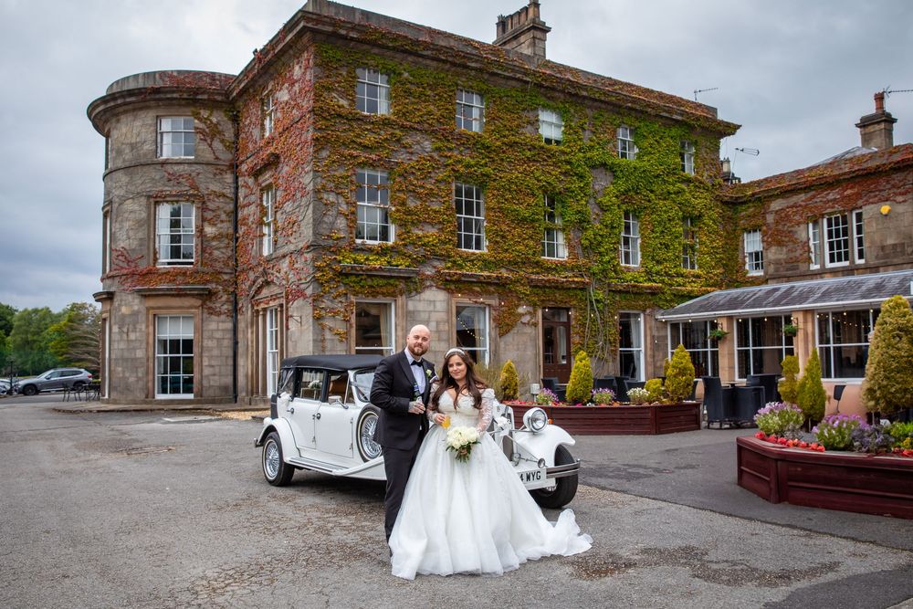 Bride and groom pose beside a vintage white car in front of an ivy-covered country house venue.