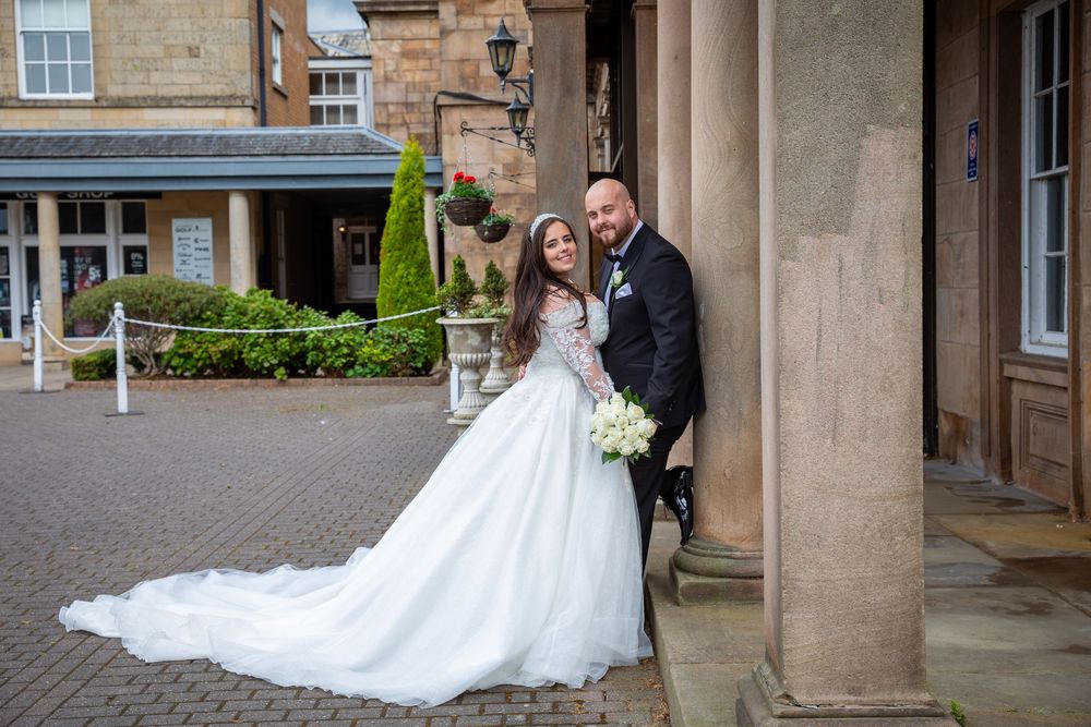 Bride and groom pose together by stone columns at the entrance of an elegant sandstone wedding venue.