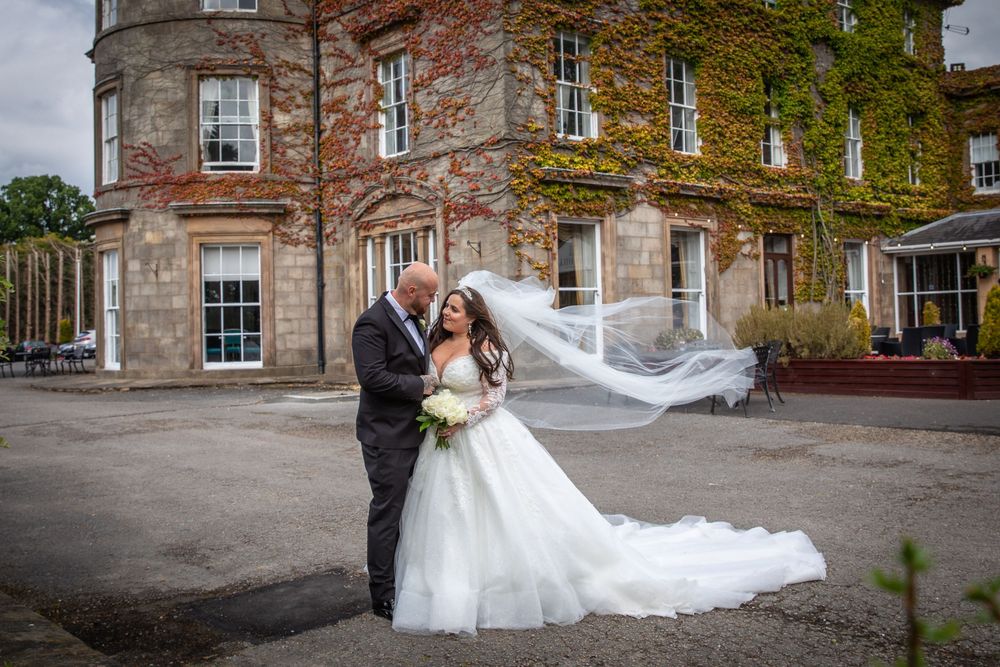 Bride's long veil blows dramatically in the wind as couple embraces outside ivy-covered country house venue.