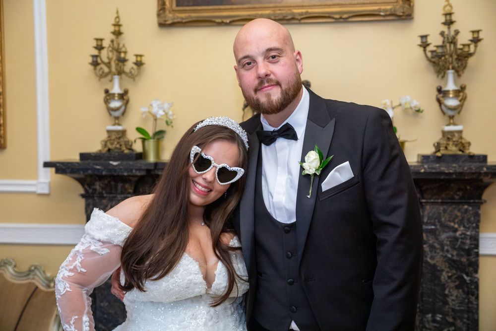Smiling bride wearing novelty heart-shaped sunglasses poses with bearded groom in tuxedo at wedding reception.