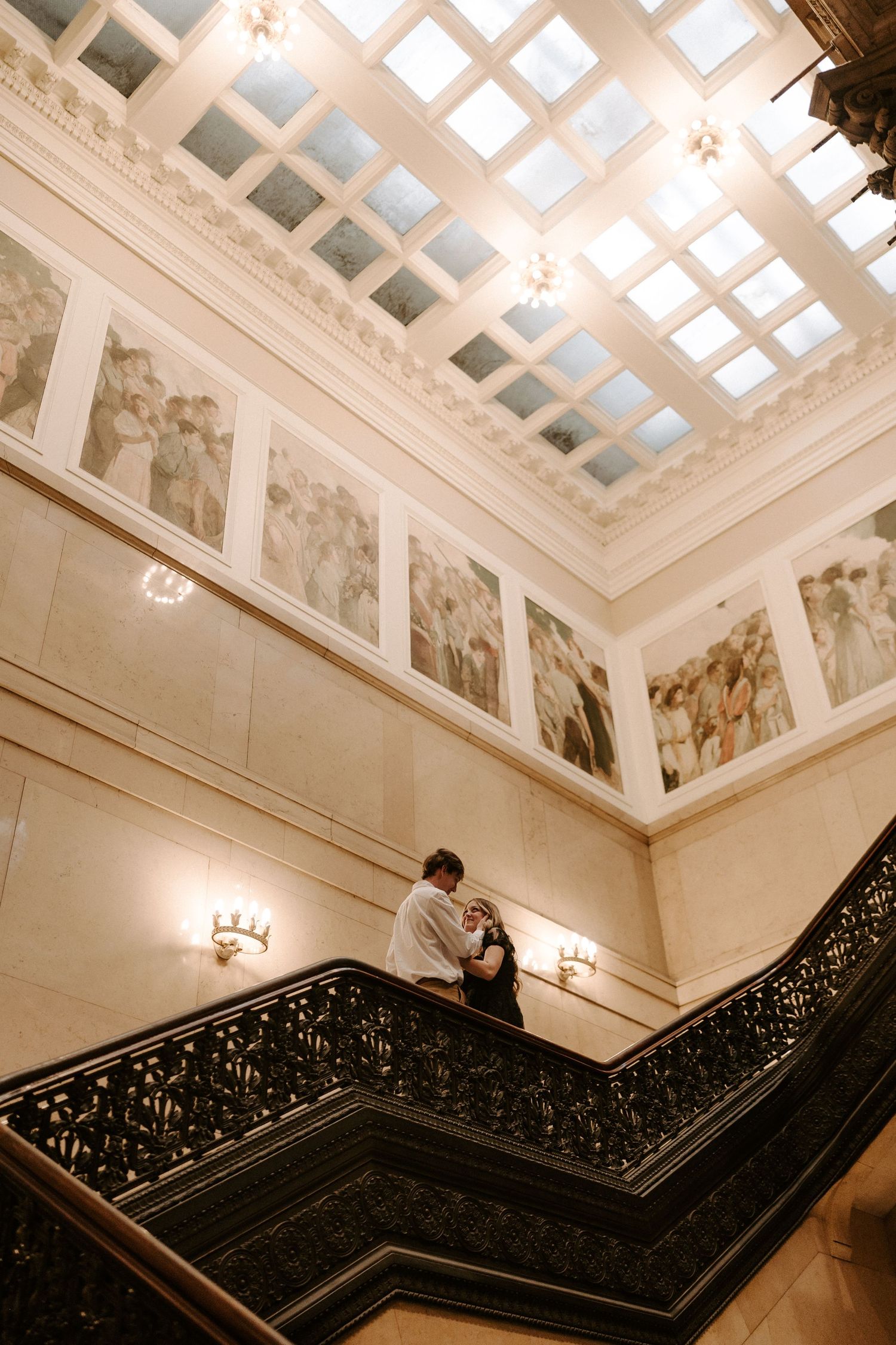 Couple embracing on ornate iron staircase beneath coffered skylight and classical murals in grand hall.