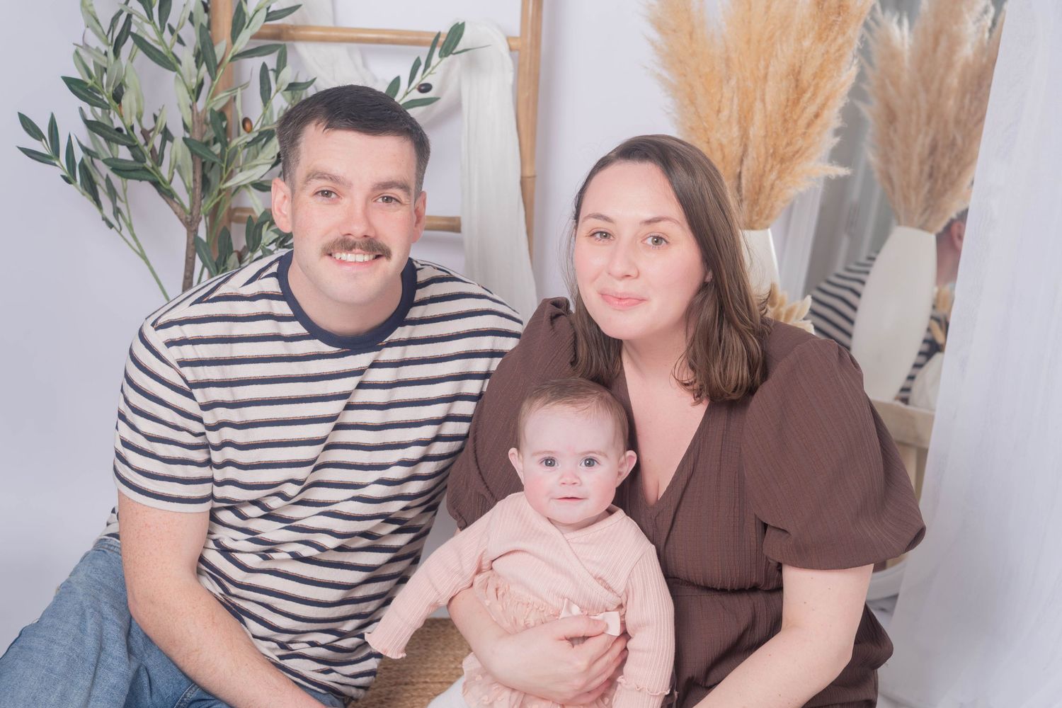 Young family portrait with father, mother, and baby sitting together in cozy studio setting. Family portrait session by K Lewis Photography