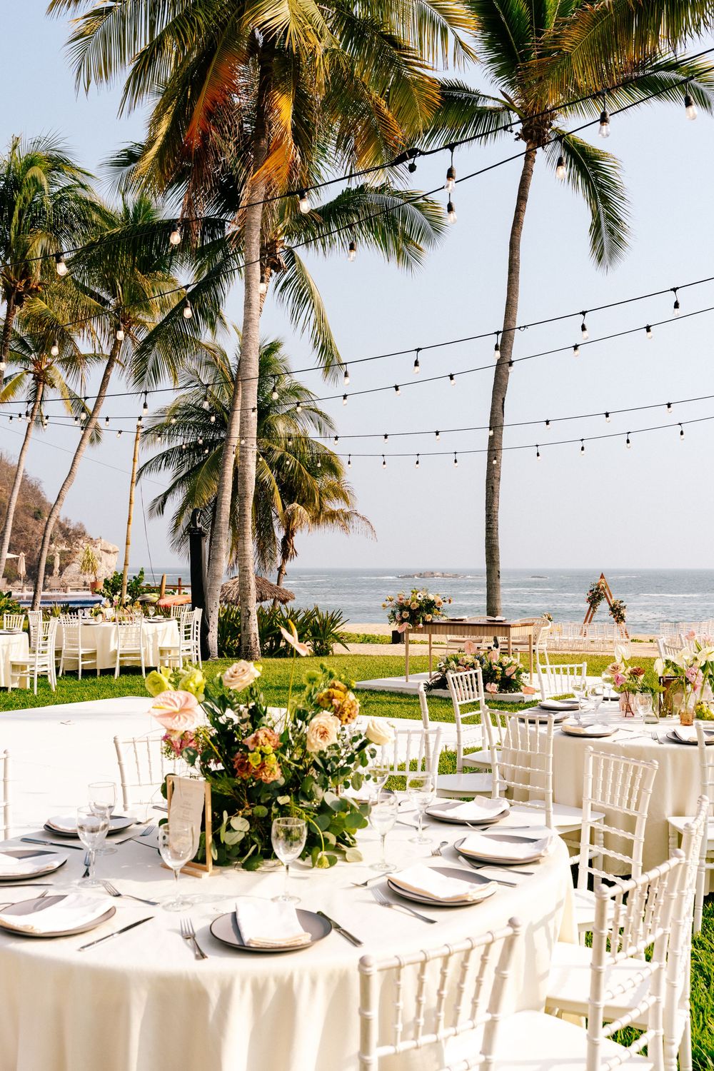 Elegant beachfront wedding reception setup with white tables, string lights and palm trees overlooking the ocean.