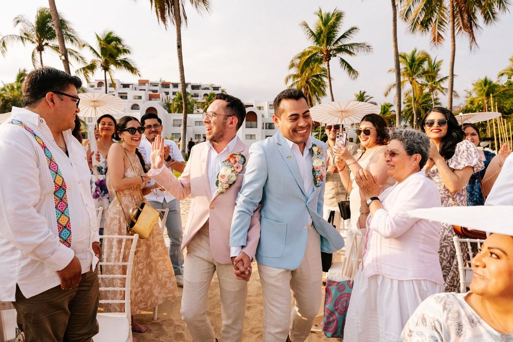 Wedding guests gather on a tropical beach with palm trees during a destination wedding celebration at sunset.