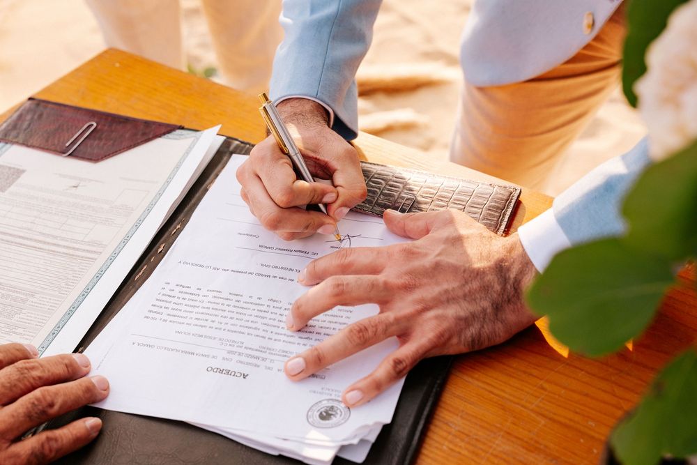 Hands signing legal documents on a wooden table with green plant leaves visible in the frame.