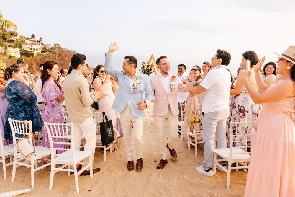 Wedding guests celebrate joyfully on a sunny beach with white chairs and pastel attire during an outdoor ceremony.