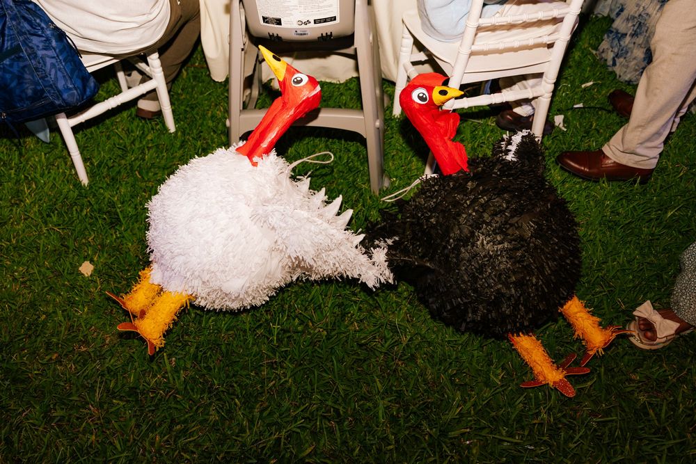 Two decorative turkey plush toys with red necks and orange feet, one white and one black, displayed on grass near lawn chairs.