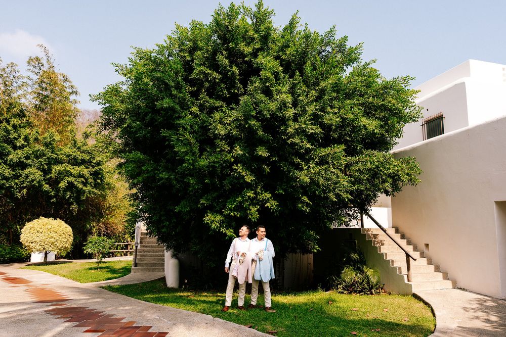 Two figures in white stand beneath a large green tree next to a modern white building on a sunny day.