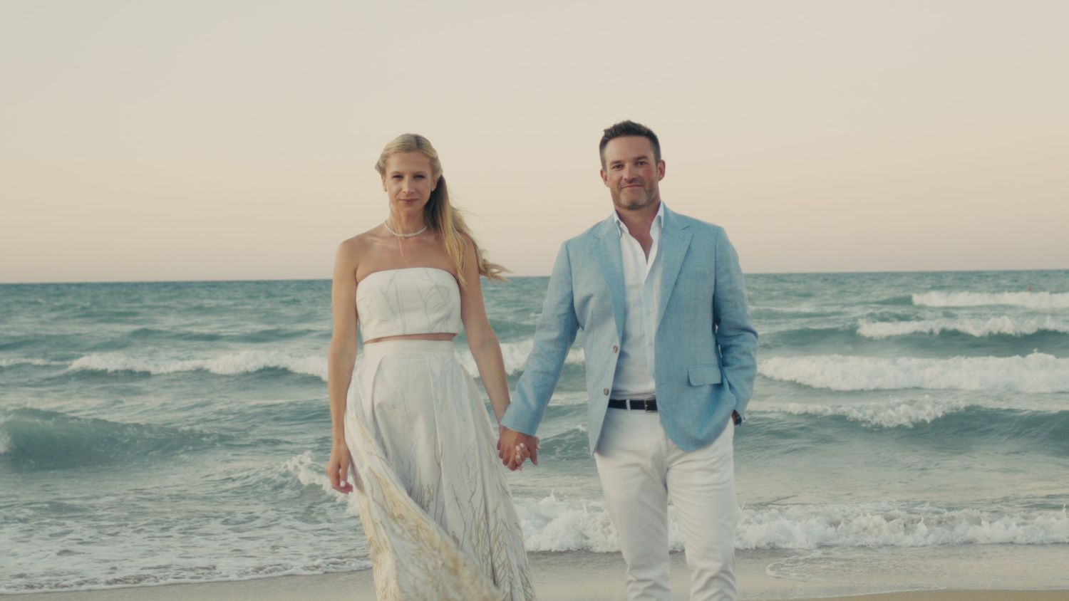 Couple holding hands walking on a sandy beach at sunset, woman in white dress, man in light blue blazer.