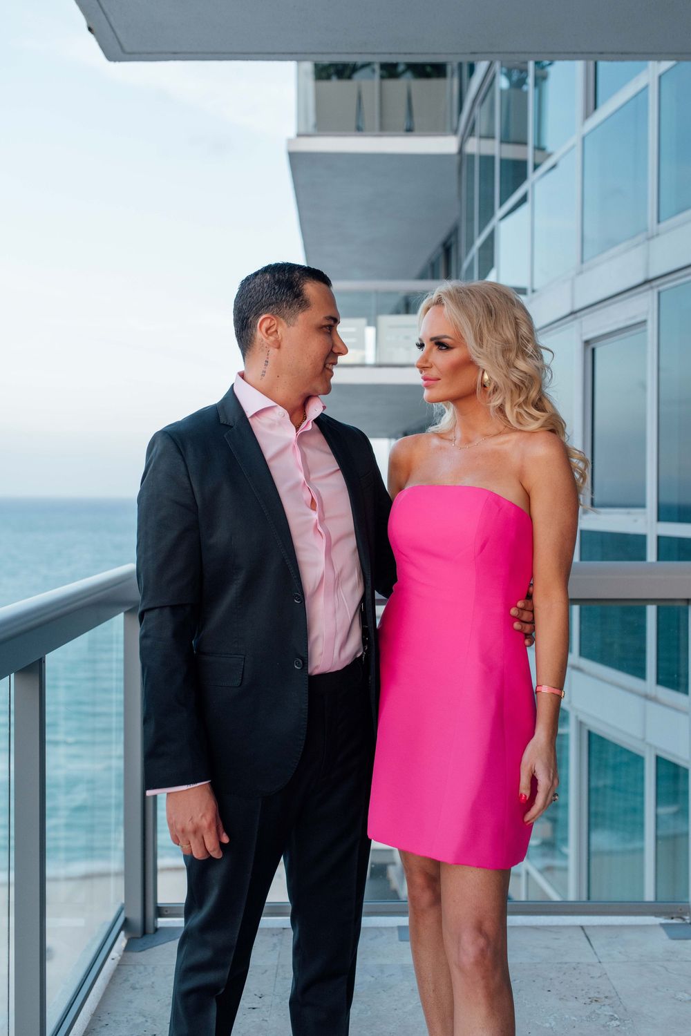Couple on a balcony, man in black suit and woman in pink strapless dress, with ocean views.