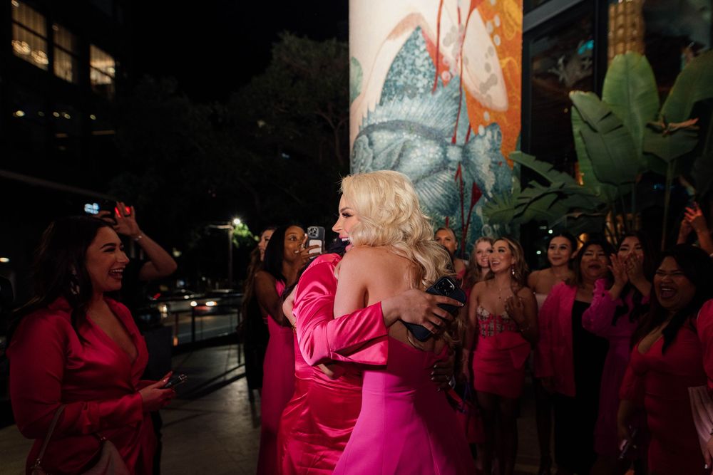Two women hugging at a pink-themed bachelorette party with guests in background at night.