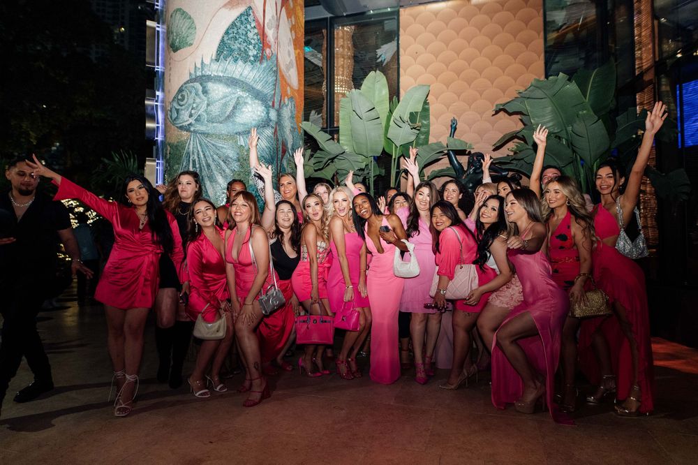 Large group of women in pink outfits posing together at a tropical-themed outdoor venue.