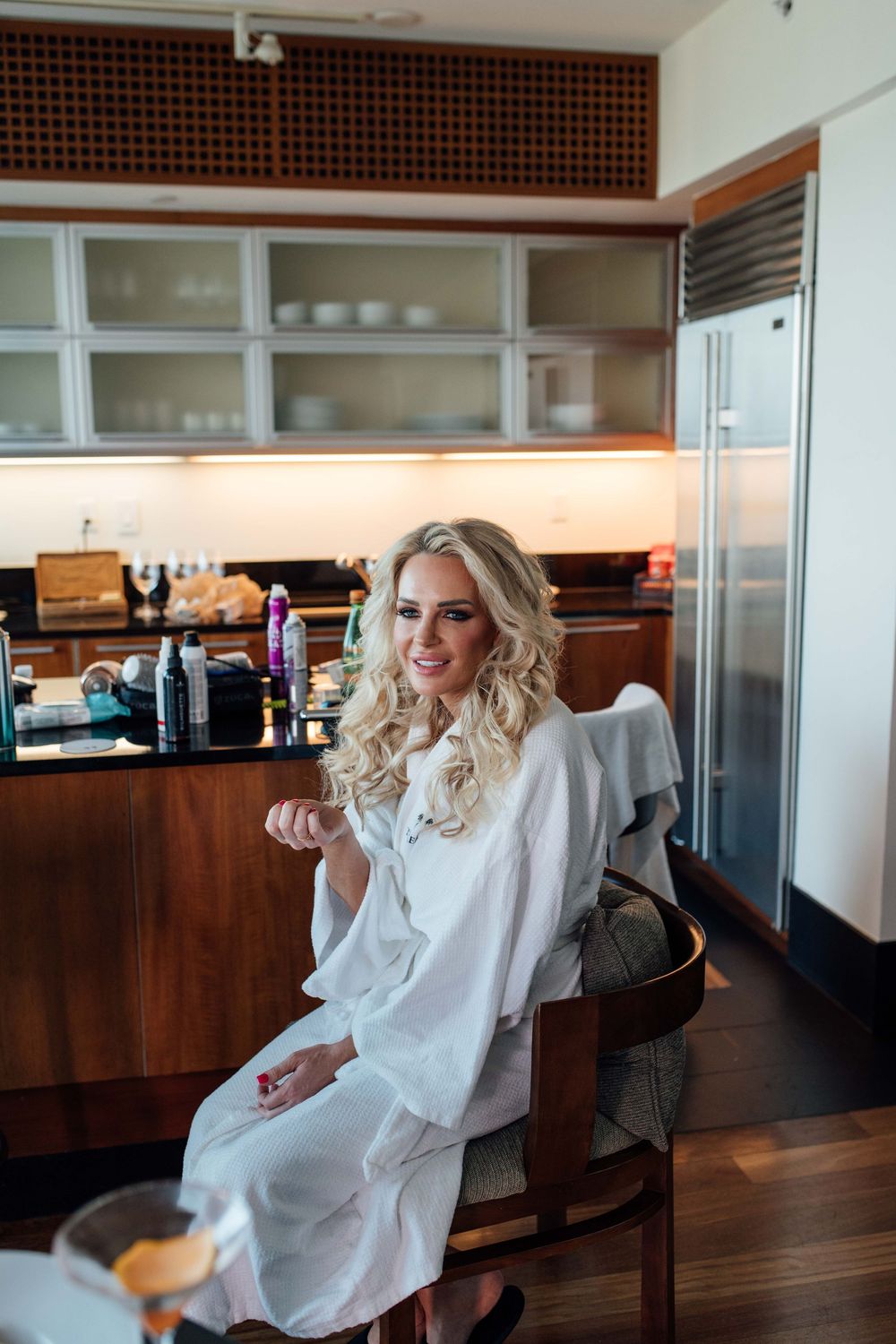 Blonde woman in white robe sitting in a modern kitchen smiling during bridal preparations.