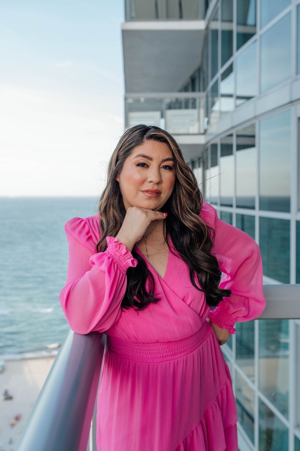 Woman in a pink chiffon dress posing on a high-rise balcony overlooking the ocean.
