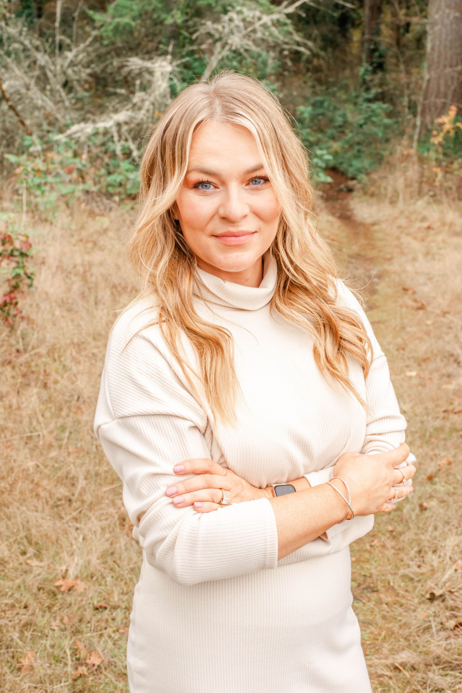 A person wearing a cream colored sweater poses outdoors in a natural setting with autumn foliage in the background.