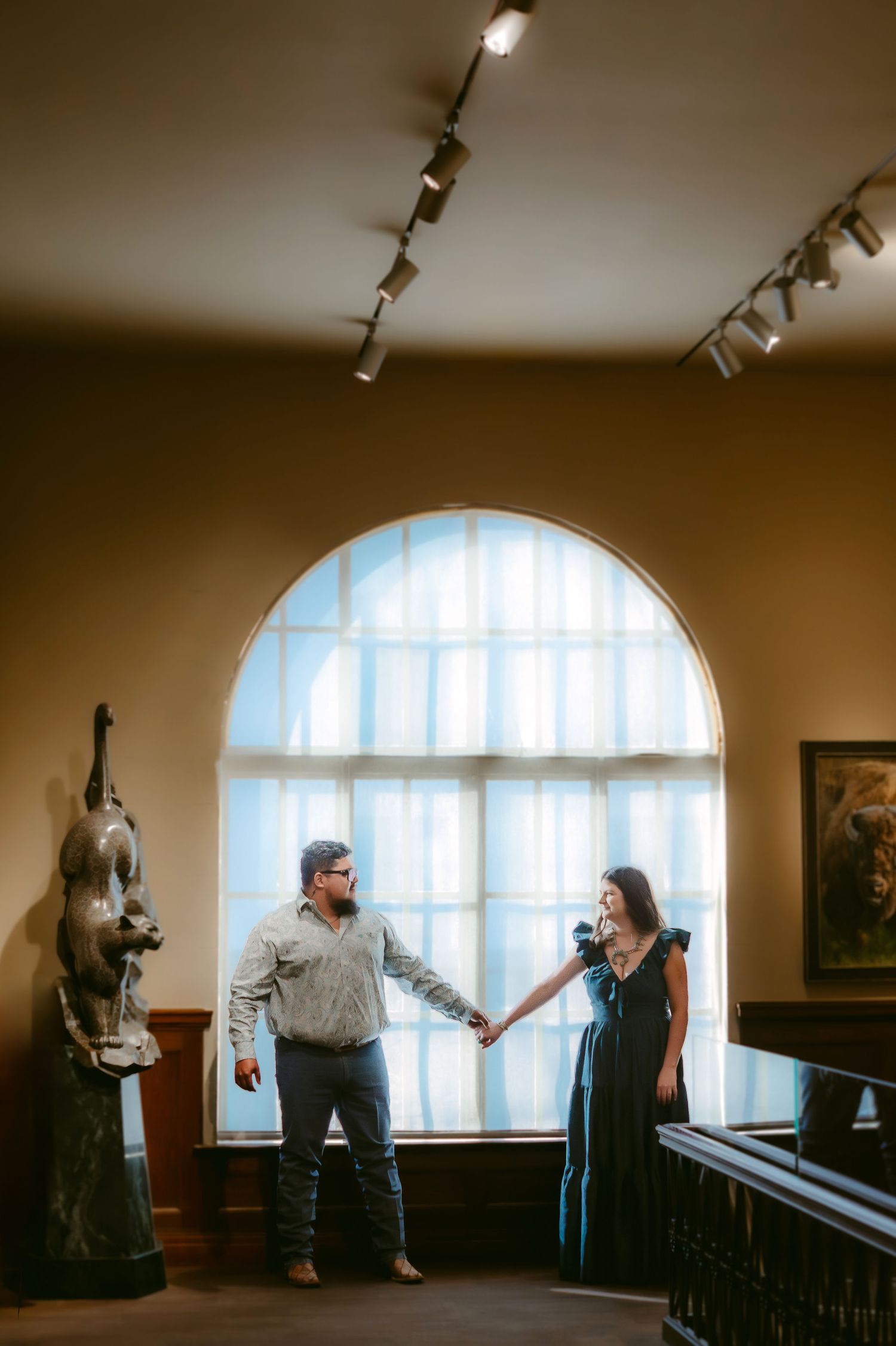 Two people engage in an animated conversation by a large arched window in a dimly lit room with track lighting.