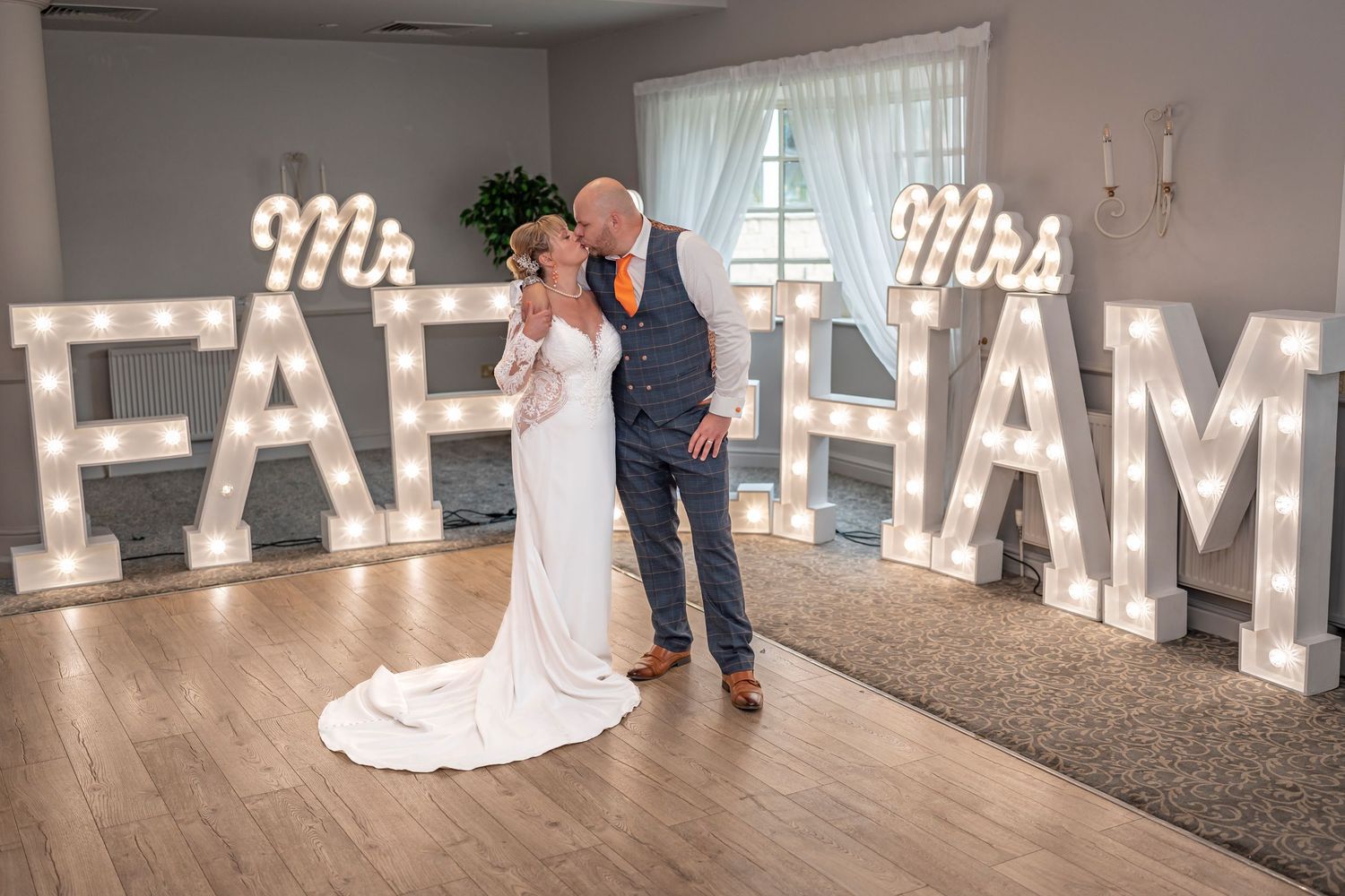 Wedding couple poses in front of illuminated marquee letters spelling their name.