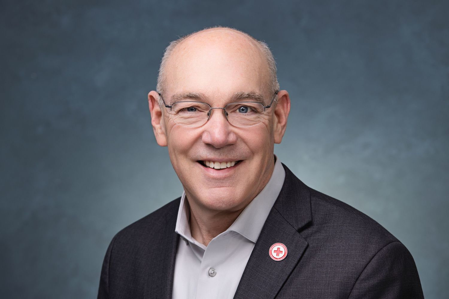 Professional headshot portrait of someone wearing a dark suit jacket and white shirt against a blue-gray background.
