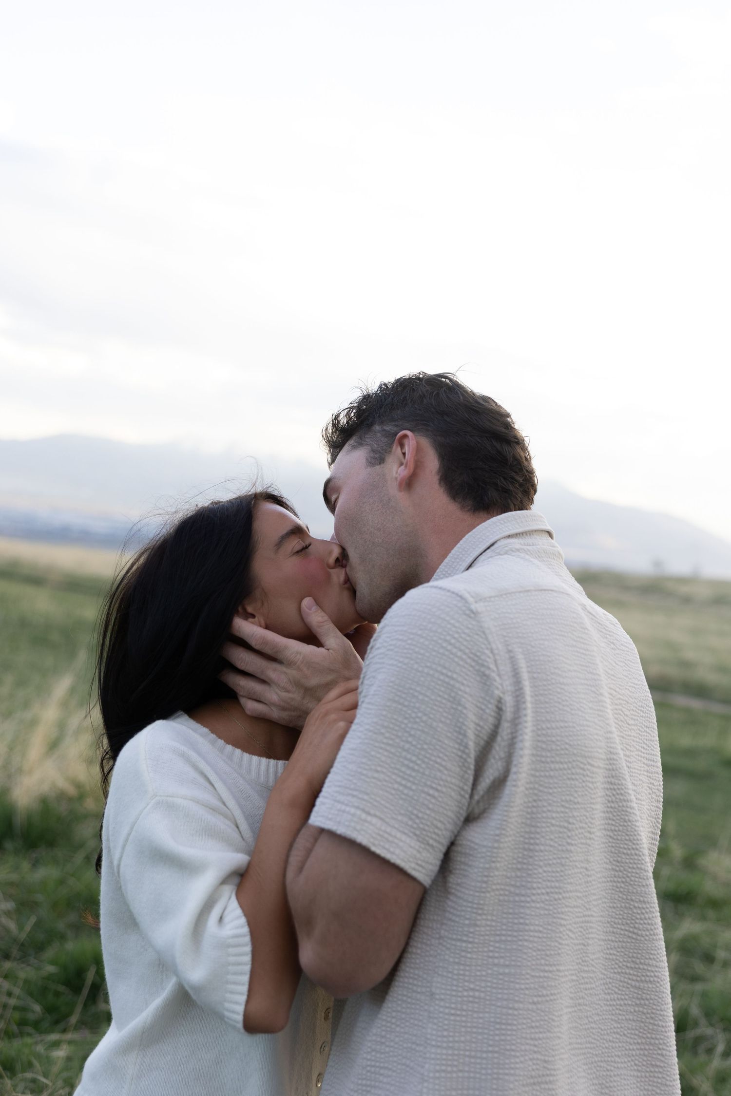 A romantic couple shares a tender kiss in a field with mountains visible in the background.