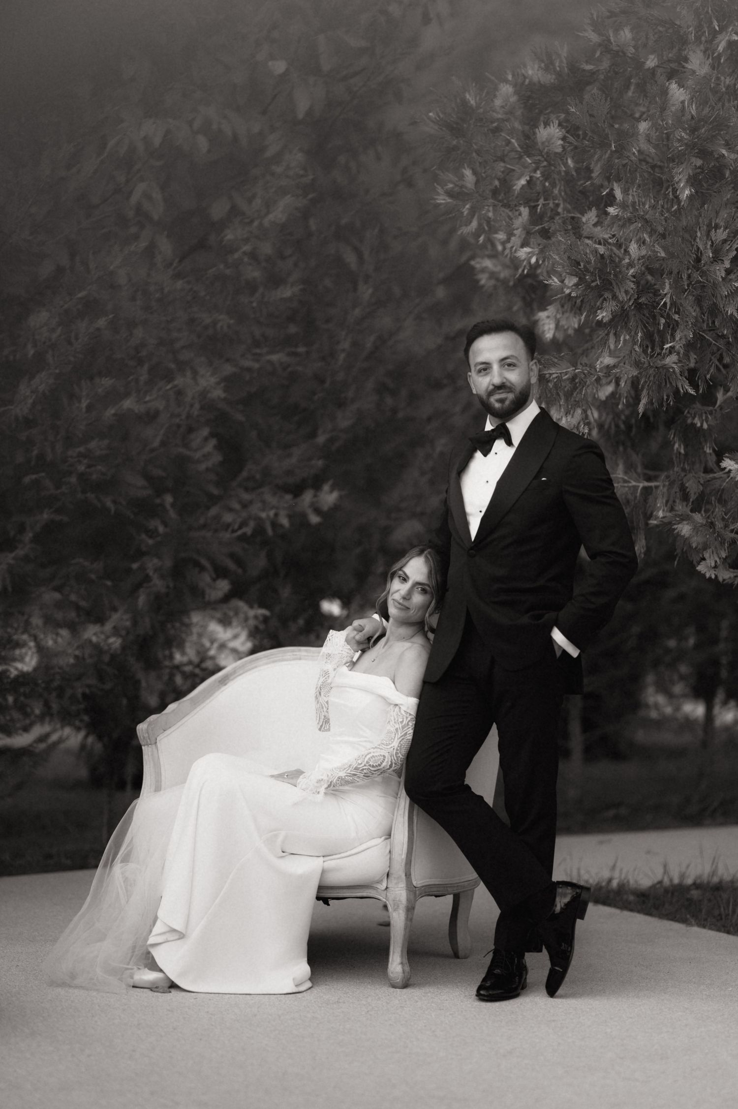 Black and white wedding portrait of bride in white gown seated in ornate chair beside groom in tuxedo outdoors.