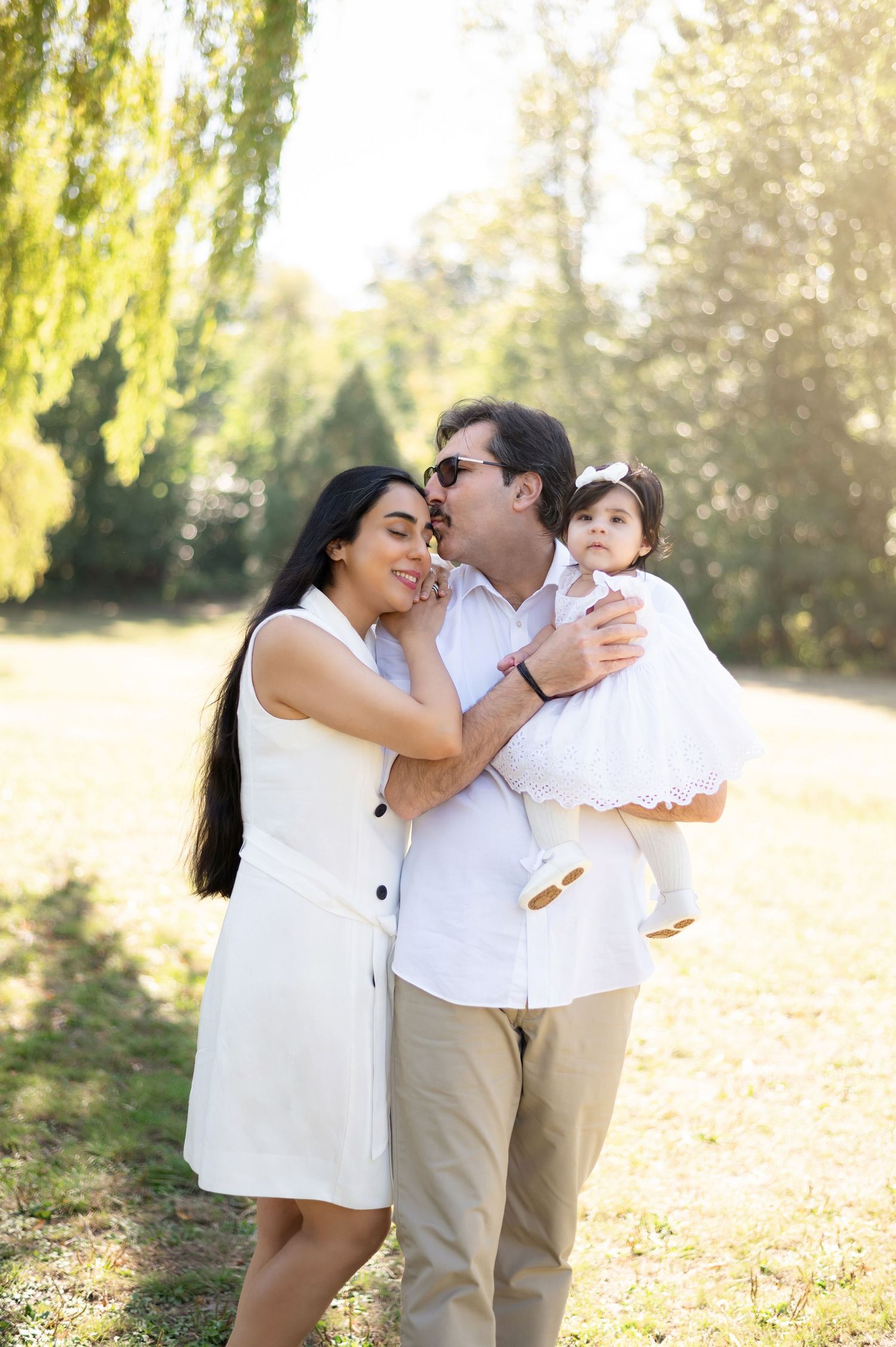 Smiling family of three with parents and baby girl posing in sunny park with willow trees behind them.
