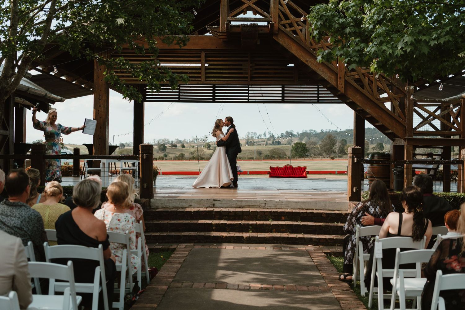 Couple sharing first kiss at outdoor wedding ceremony under rustic wooden pergola with guests seated in white chairs.