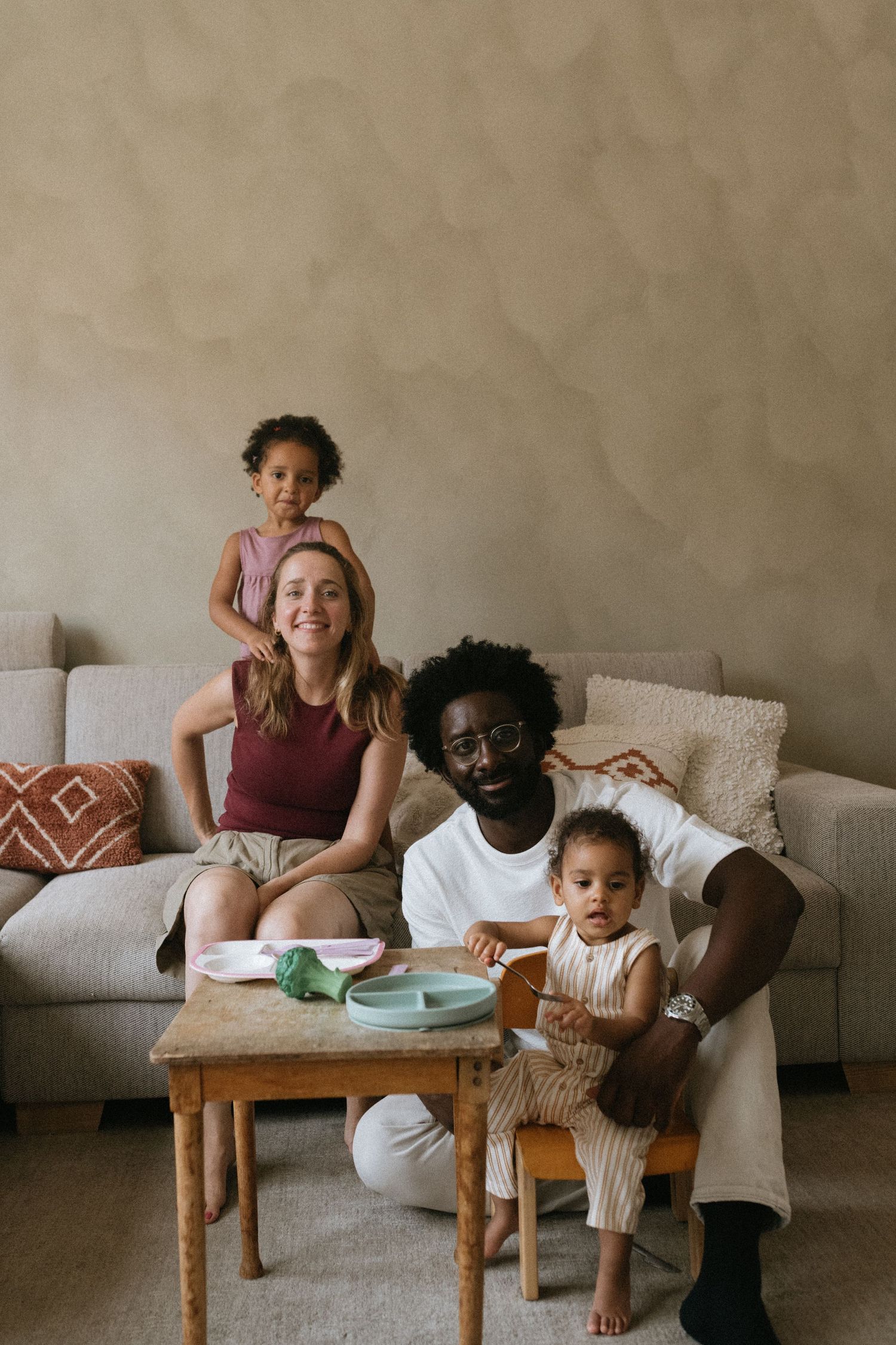 Multiracial family of four posing together in a cozy living room with a sofa, small wooden table, and decorative pillows.