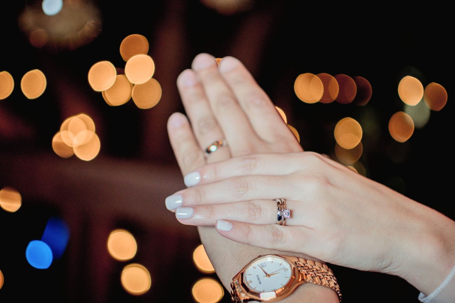 Wedding rings sparkle against blurred bokeh lights with elegant watch details.
