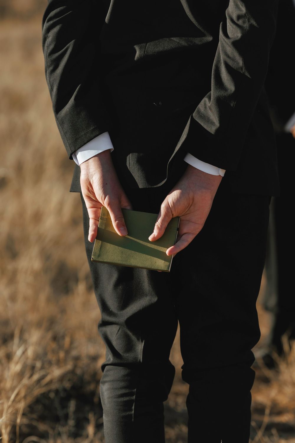 Close-up of groom in black suit holding small green book during outdoor wedding ceremony.