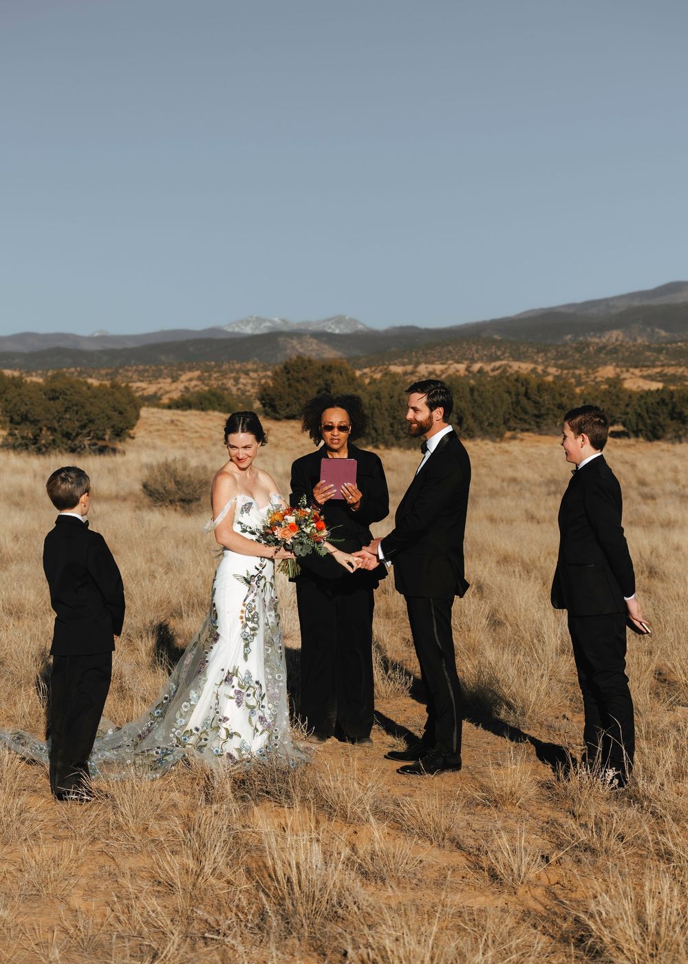 Wedding ceremony in golden field with bride in floral gown, groom, officiant, and two attendants.