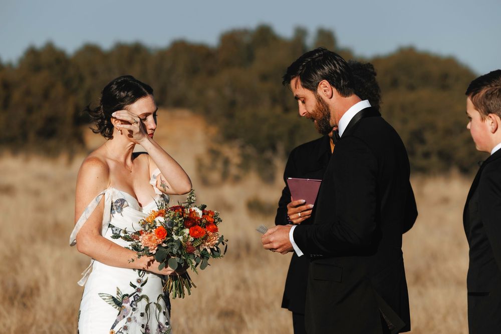 Emotional bride wiping tear during outdoor wedding ceremony while groom reads vows in open field.