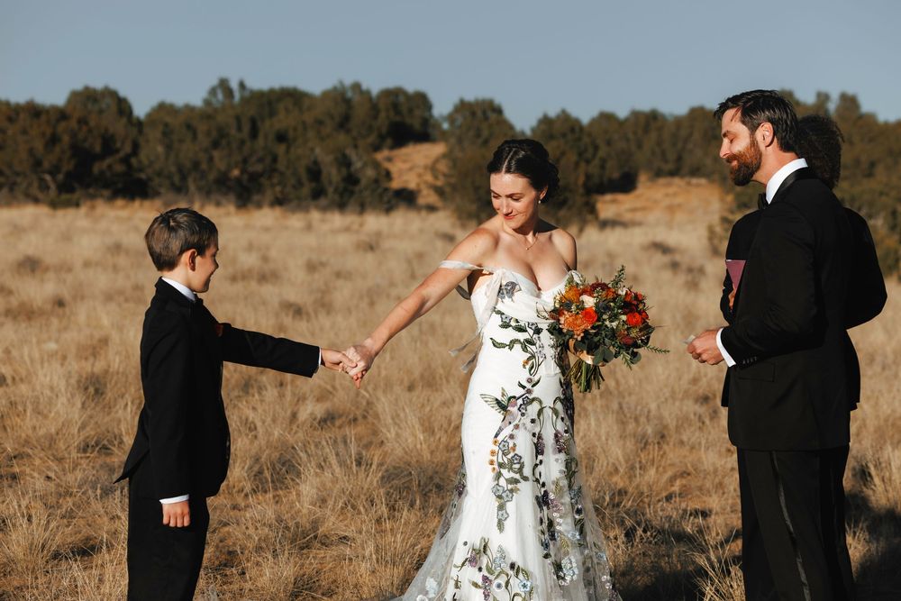 Bride in floral gown holds hands with young boy in black suit at outdoor wedding ceremony in golden field.
