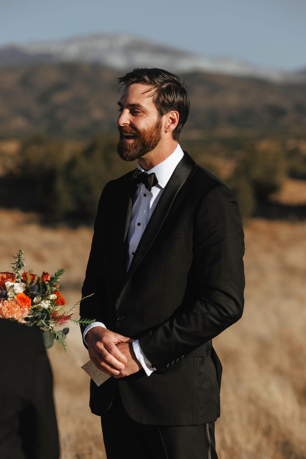 Bearded groom in black tuxedo smiles during outdoor wedding ceremony with mountain backdrop.