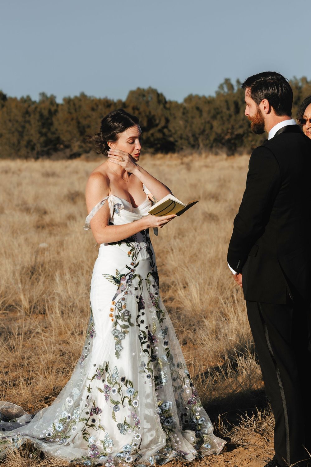 Emotional bride in floral wedding gown reads vows from book during outdoor field ceremony.