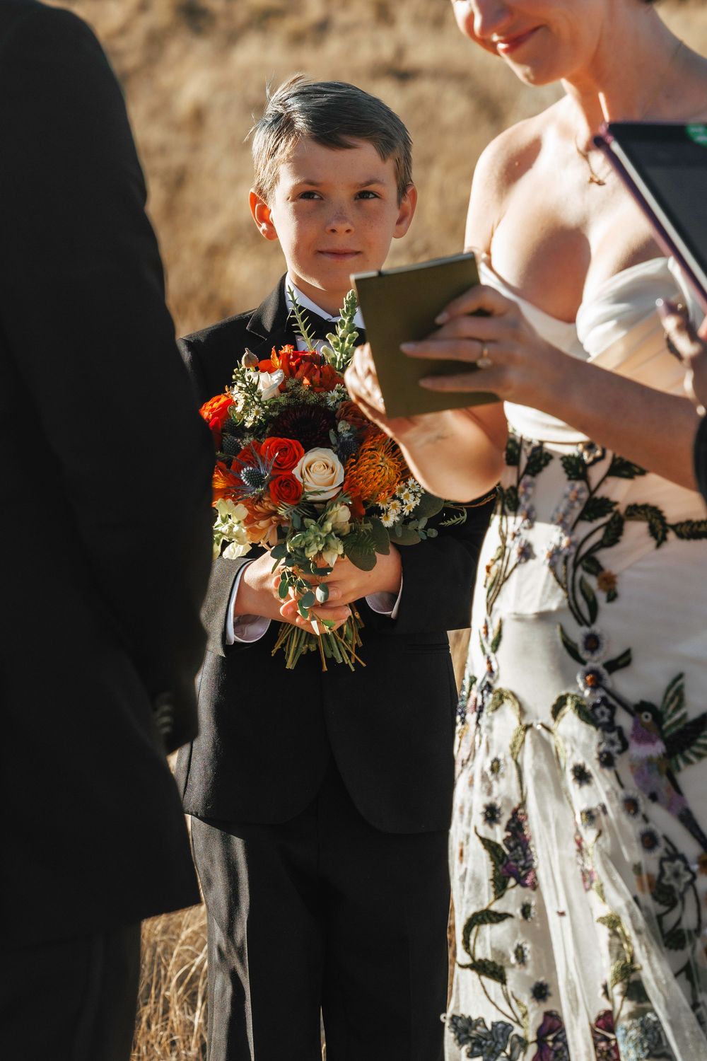 Young ring bearer in black suit holds colorful orange and white bouquet during wedding ceremony.