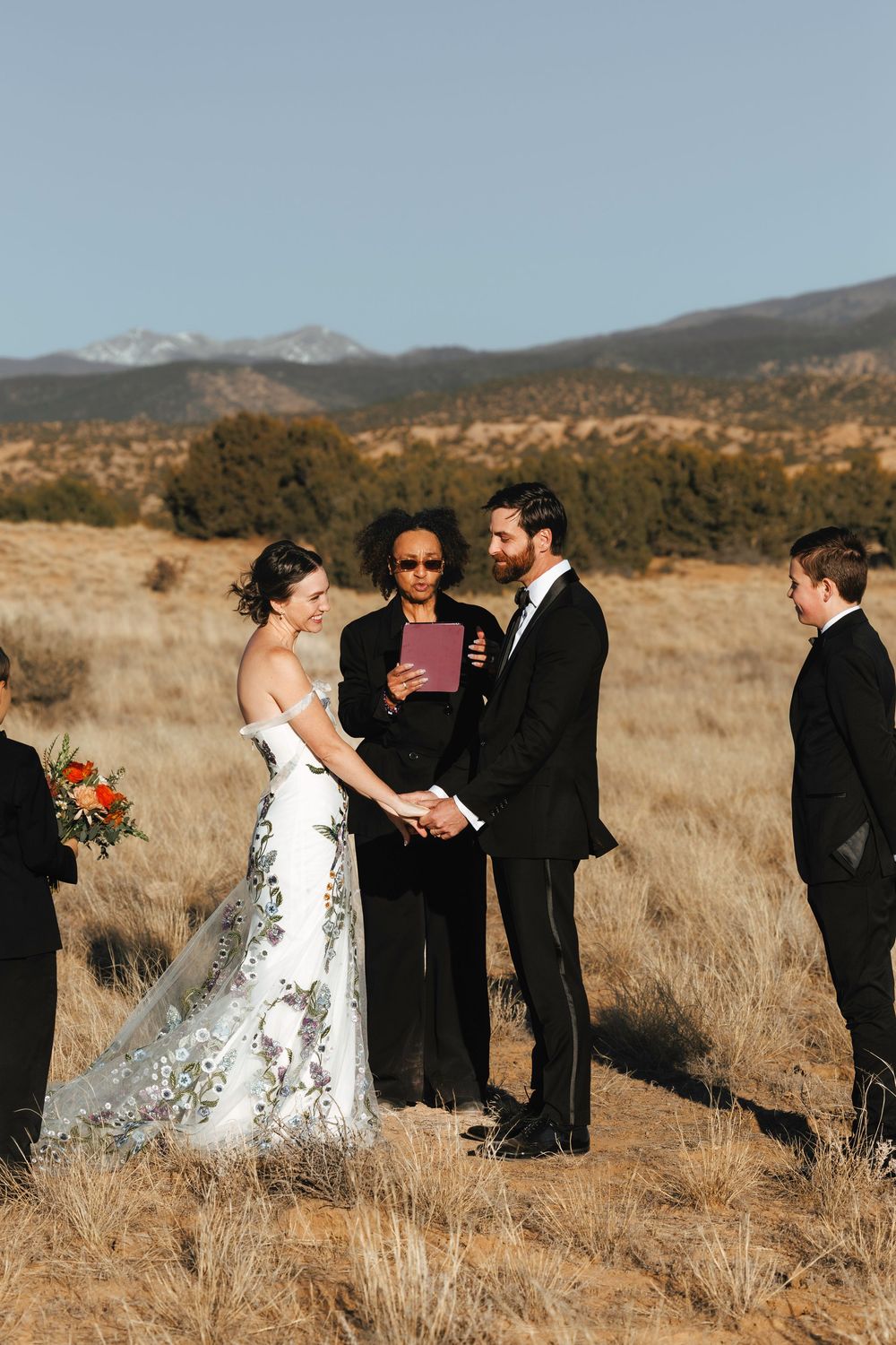 Bride and groom exchange vows with officiant in golden field with snow-capped mountains behind.