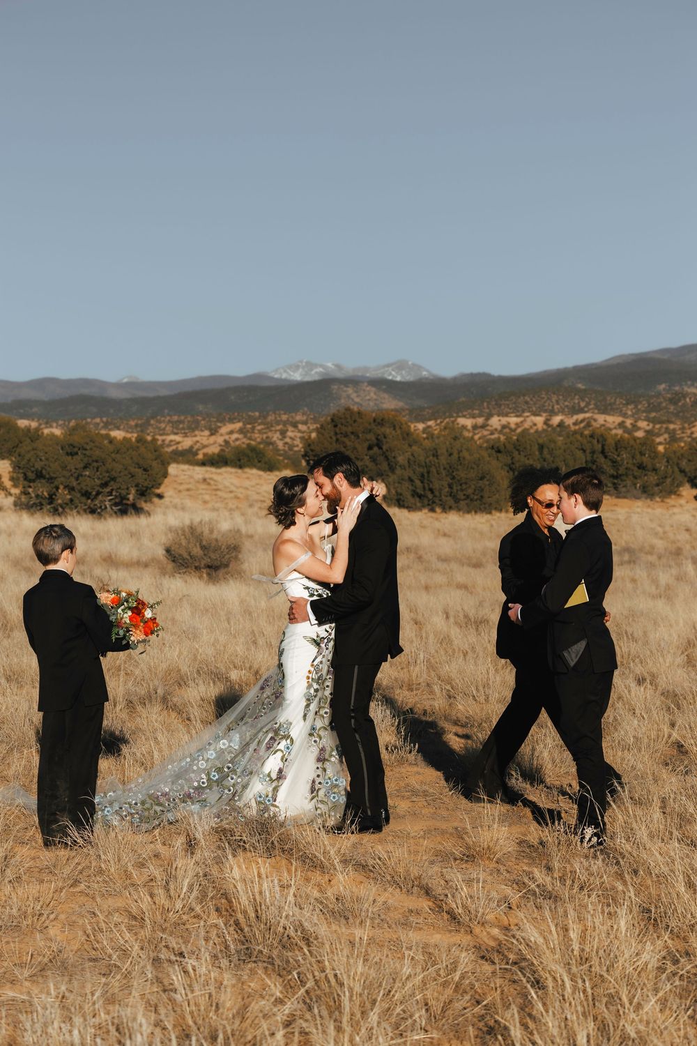 Newlyweds share first kiss in golden field as officiant and ring bearer celebrate nearby.