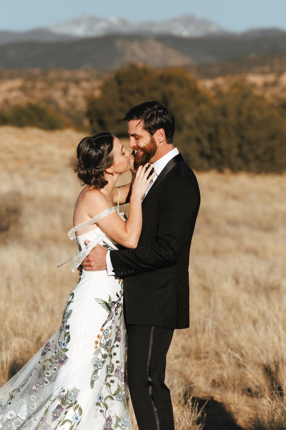 Bride and groom embrace romantically in sunlit field with mountains and evergreens in background.