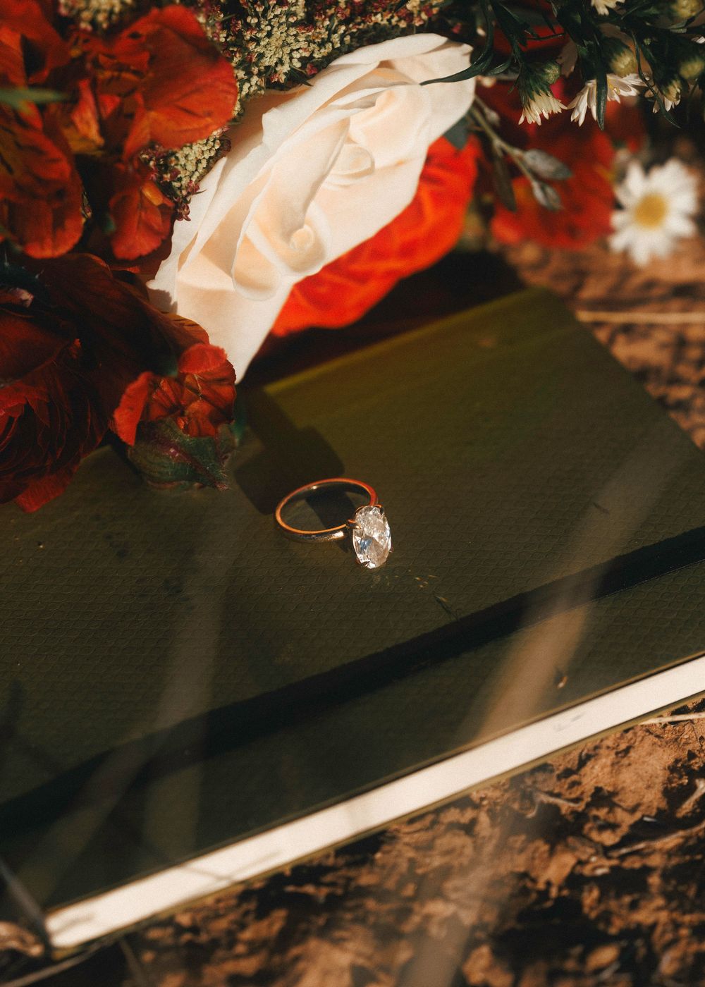 Diamond engagement ring rests on dark book beside vibrant red and white wedding bouquet.