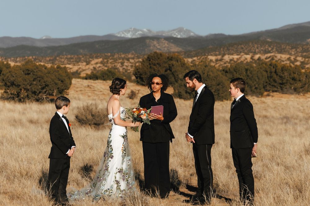 Outdoor wedding ceremony in open field with snow-capped mountains, officiant reading vows to couple.