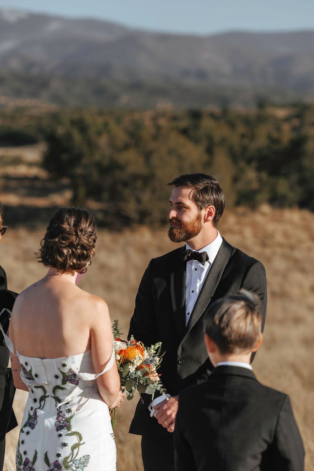 Groom in black tuxedo gazing at bride during outdoor wedding ceremony with mountain landscape.