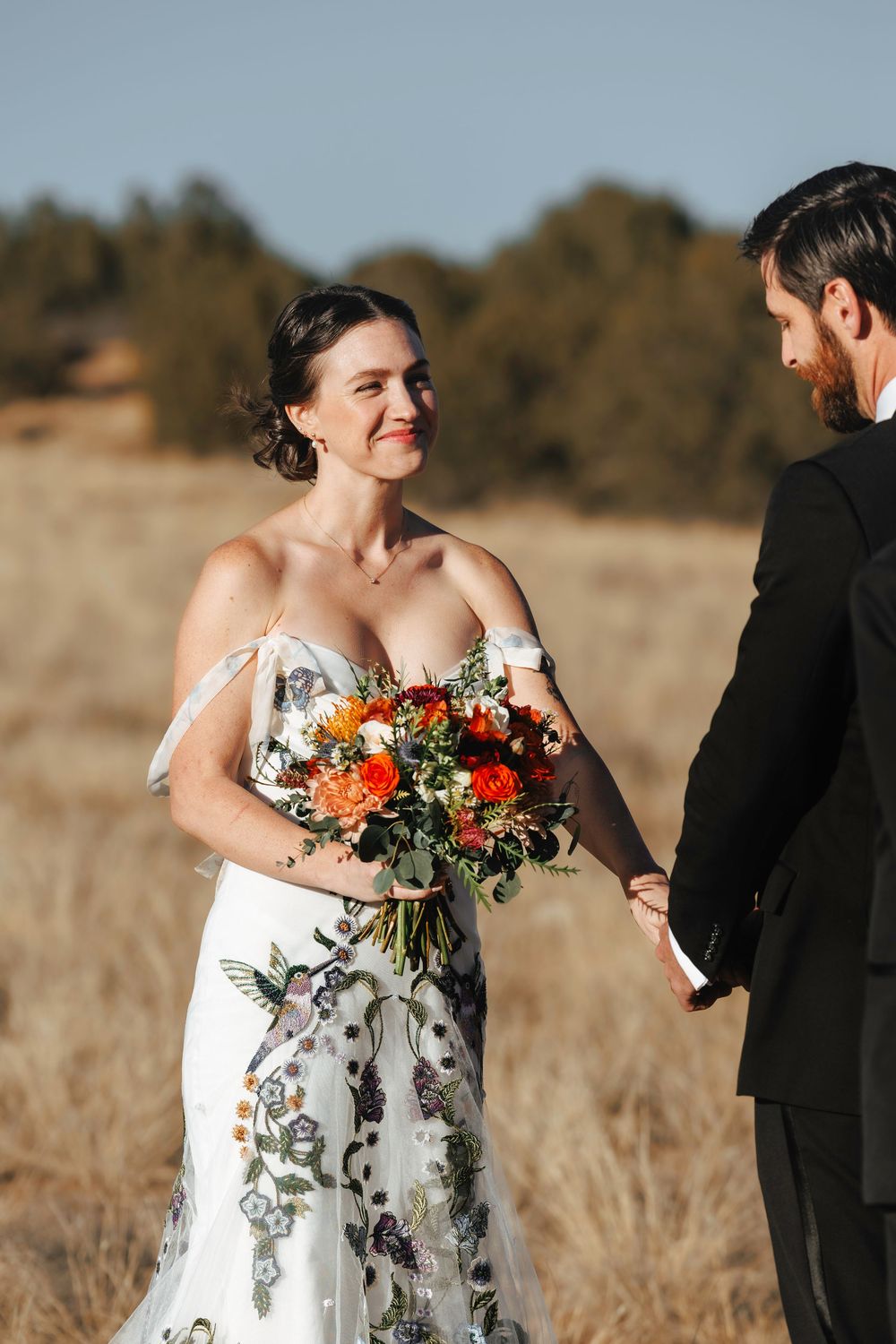 Smiling bride in floral wedding gown holding vibrant orange bouquet while exchanging vows outdoors.